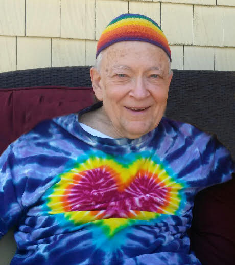 Rabbi Hanan Sills smiling, wearing a colorful knit cap and a tie-dye shirt with a heart design.